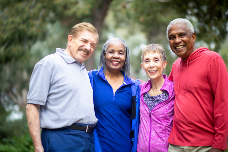 Four senior adults standing close together, smiling in bright clothing in front of a blurry forest backgdrop